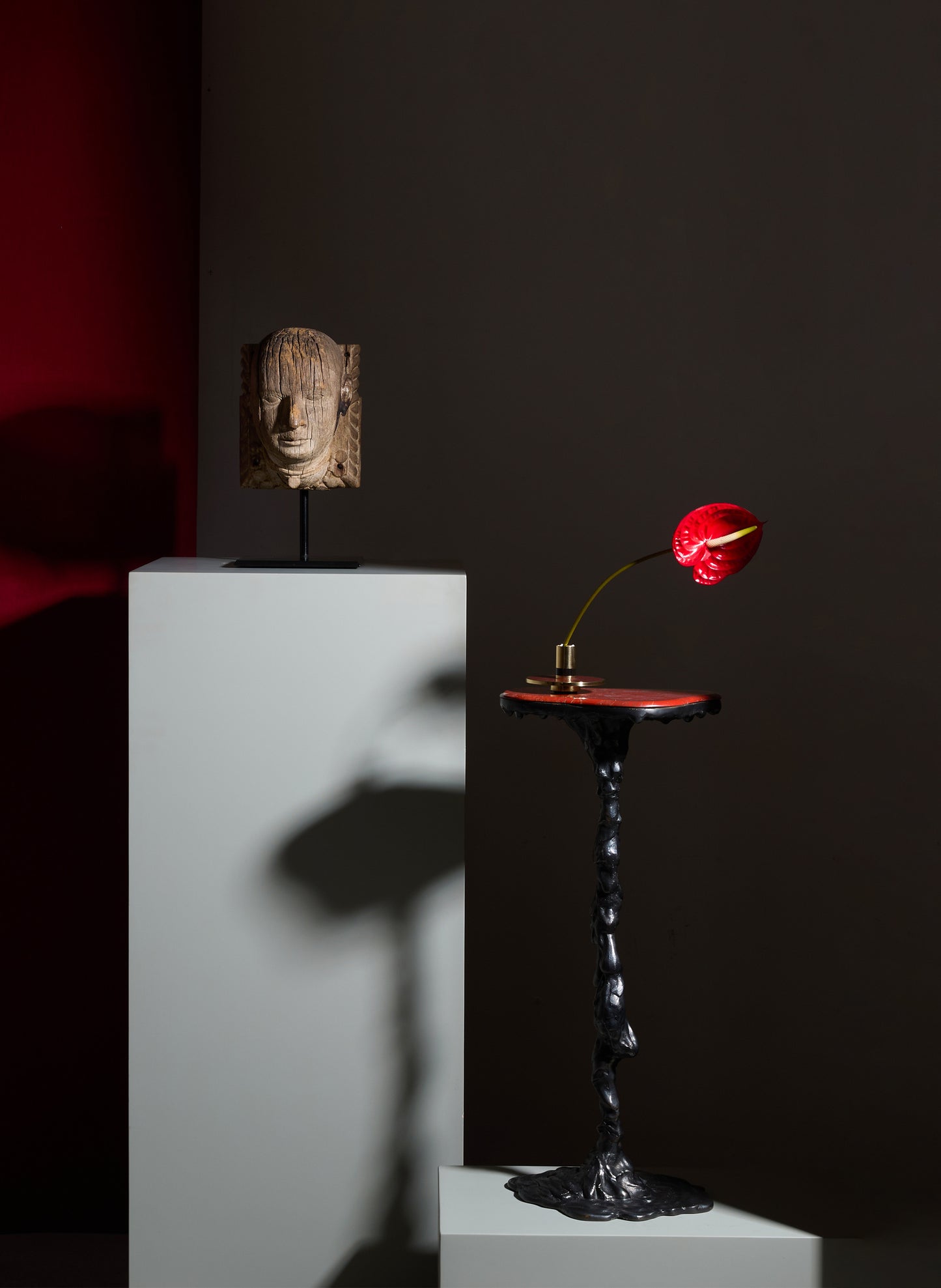 Decorative sculpture head made of carved wood, mounted on a black stand, with a textured surface and neutral tones. A red anthurium flower with a curved yellow spadix on a black decorative pedestal with a twisted, sculptural base, set against a dark background.