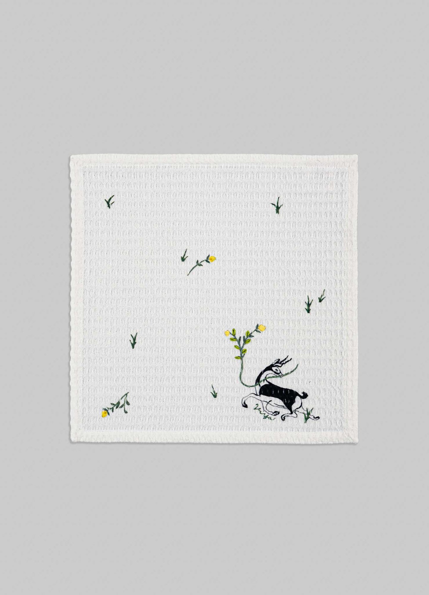 A white embroidered cushion cover featuring a whimsical illustration of a dragon with floral and leaf motifs, accented with yellow flowers. The fabric appears textured with a subtle waffle weave pattern, set against a plain grey background.