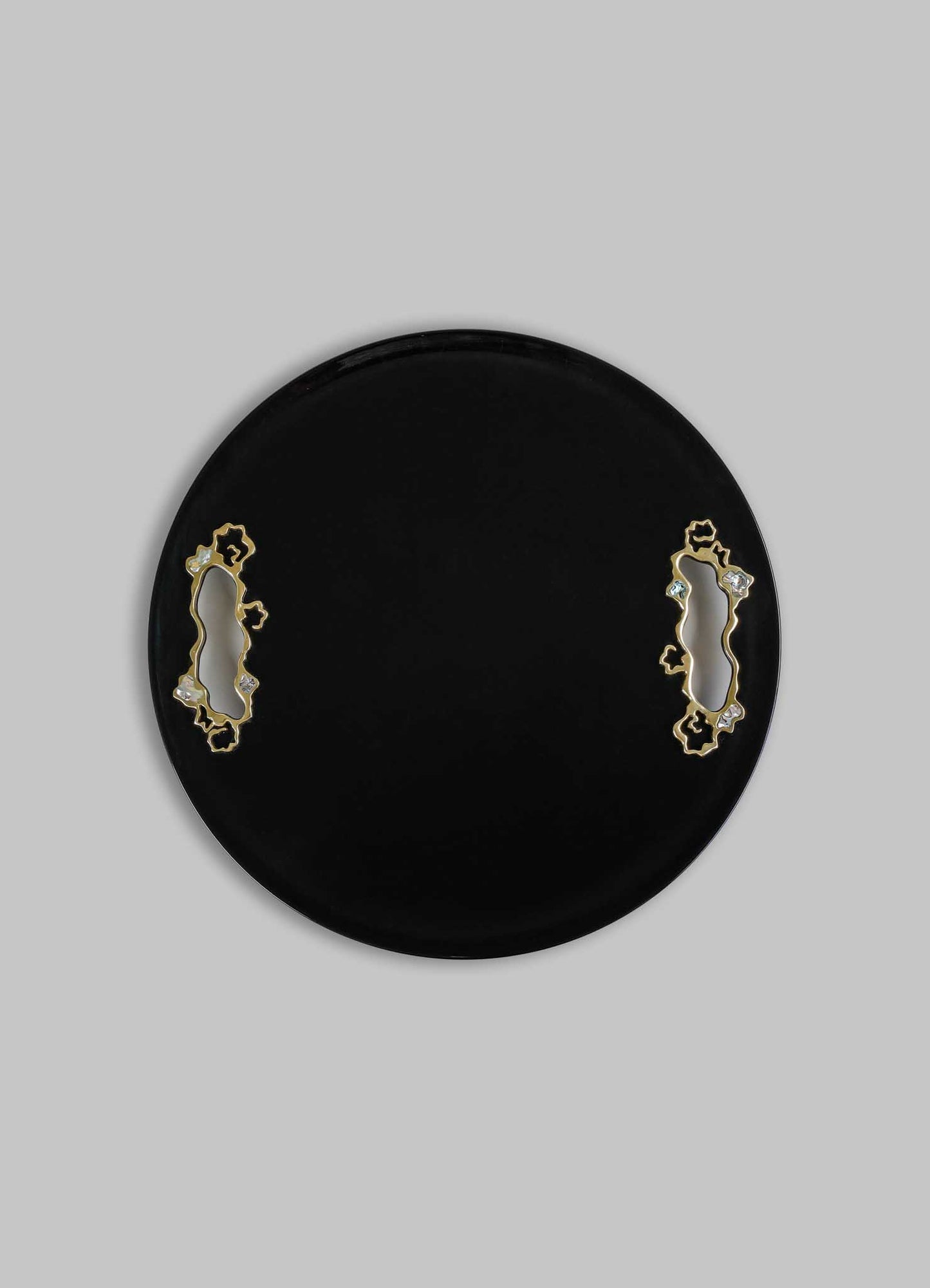 A round black tray with decorative cutouts bordered by gold-tone metal and small clear embellishments, set against a light grey background.