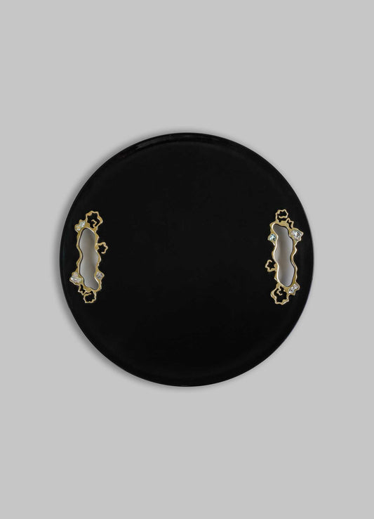 A round black tray with decorative cutouts bordered by gold-tone metal and small clear embellishments, set against a light grey background.