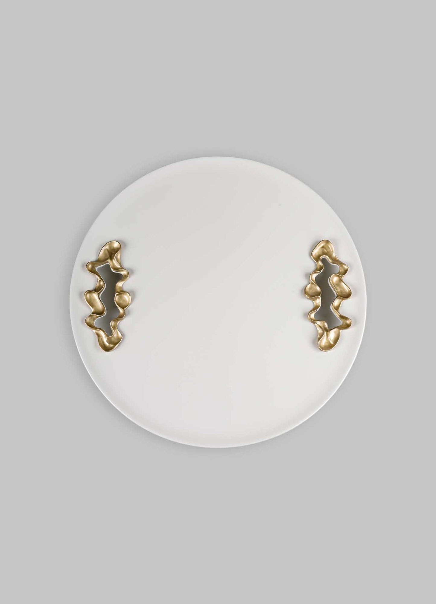 A round white ceramic tray with abstract brass handles featuring organic, irregular shapes, set against a light grey background.