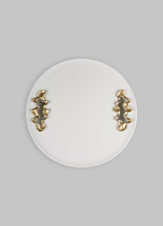 A round white ceramic tray with abstract brass handles featuring organic, irregular shapes, set against a light grey background.
