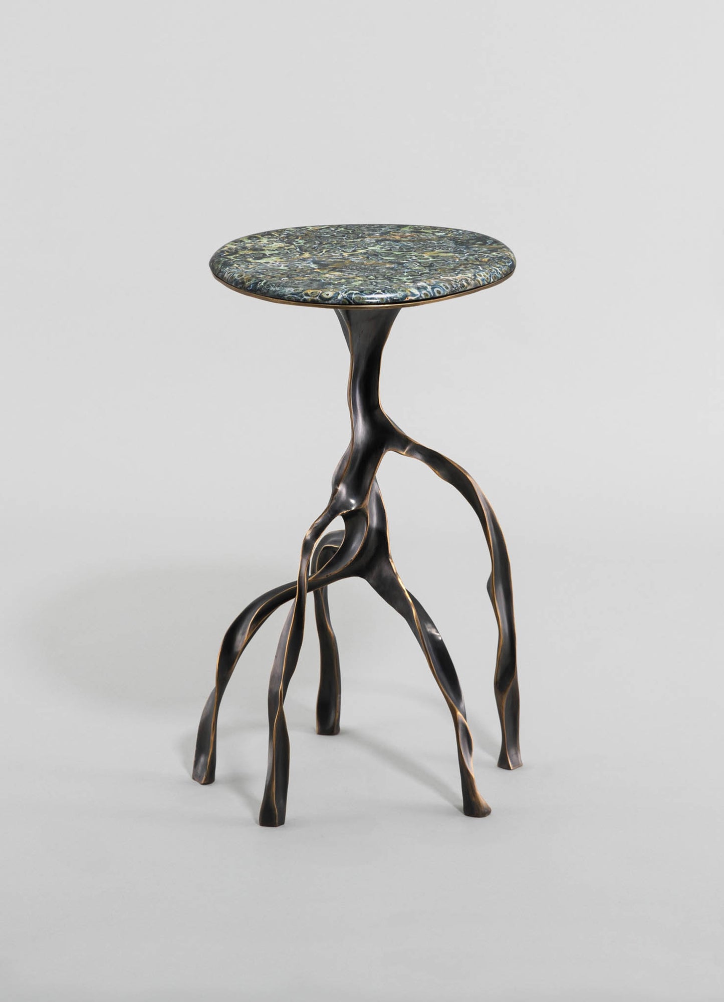 A side table with a round top made of patterned stone or marble in shades of dark and gold tones, supported by a sculptural metal base resembling intertwined branches or roots with a dark finish. The design combines organic motifs with contemporary style.