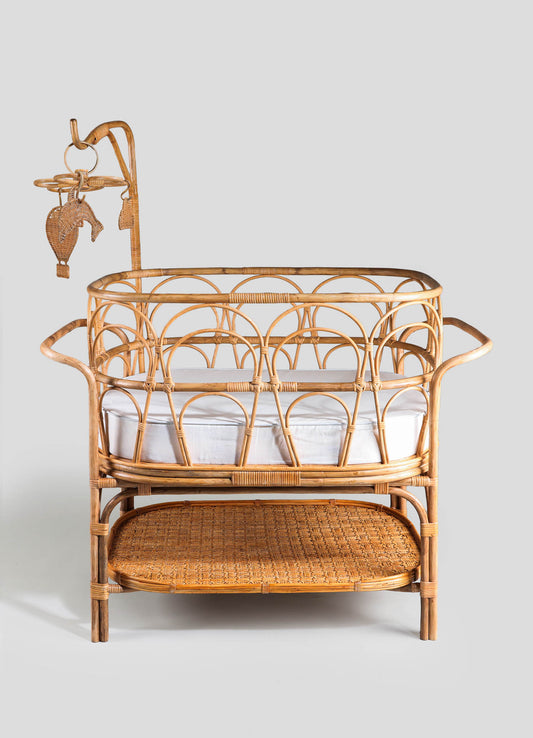 Rattan crib with an intricate curved design, featuring a natural wood finish, a white mattress, and a lower woven rattan shelf. Includes a matching mobile with hanging animal-shaped decorations, all in a handcrafted, rustic style.