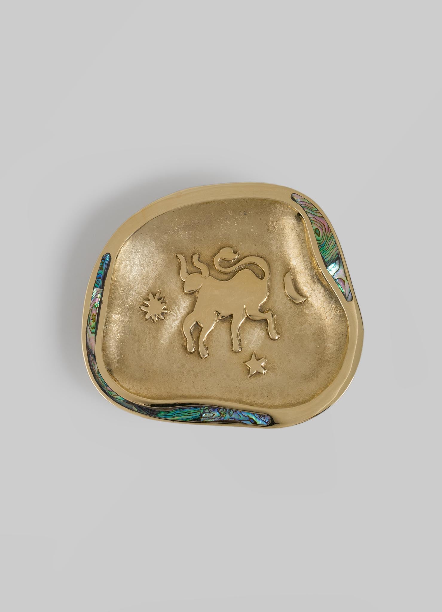 A decorative brass dish with a rounded, irregular shape, featuring an embossed lion motif and small star and sun symbols, accented with abalone shell inlay along the rim.