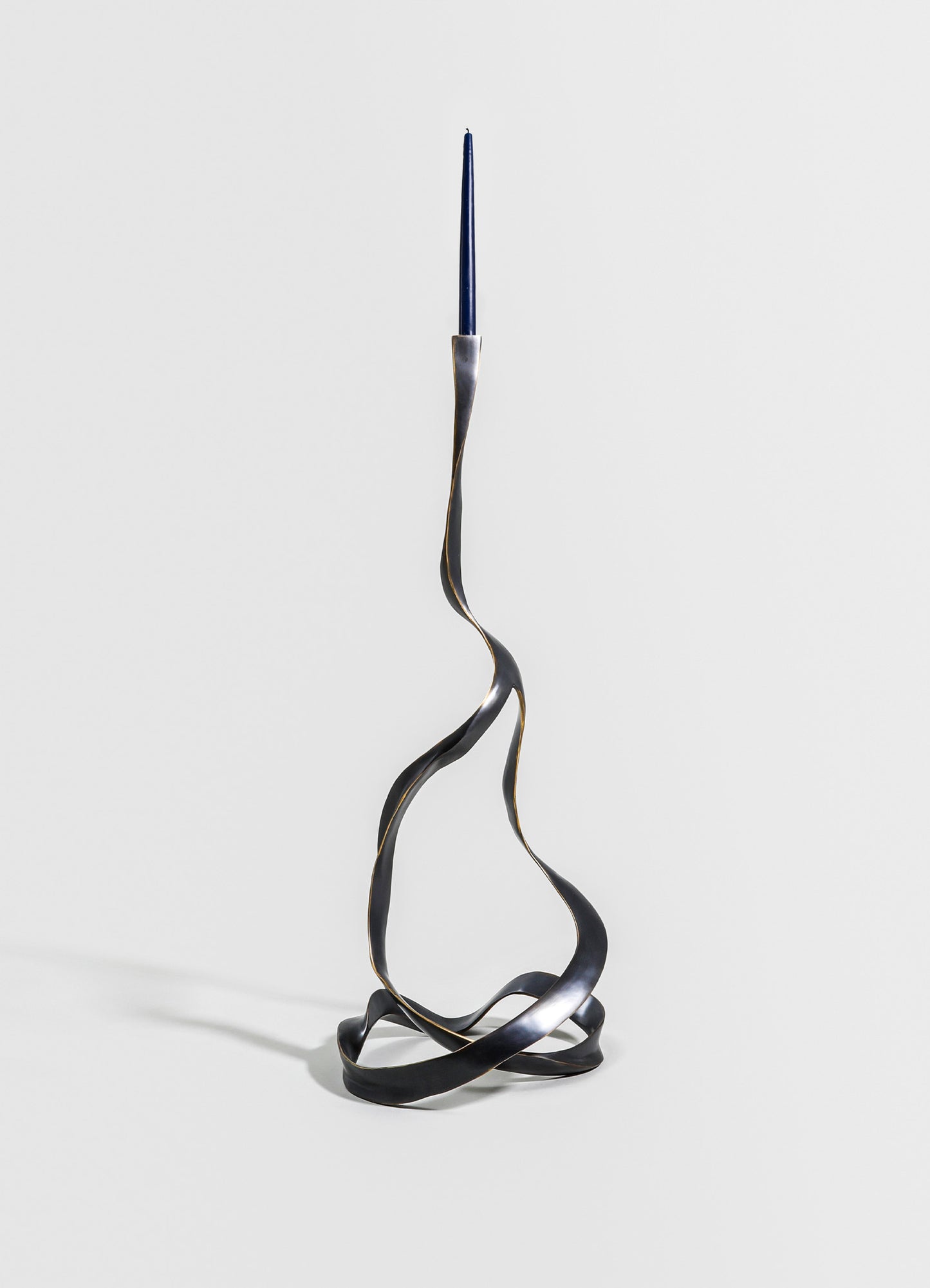 A sculptural metal candle holder with a twisted, wave-like design in a dark, matte finish, holding a tall, slender blue candle at the top. The piece has an abstract, contemporary style.