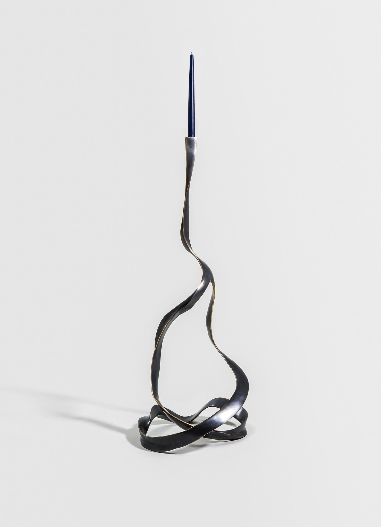 A sculptural metal candle holder with a twisted, wave-like design in a dark, matte finish, holding a tall, slender blue candle at the top. The piece has an abstract, contemporary style.