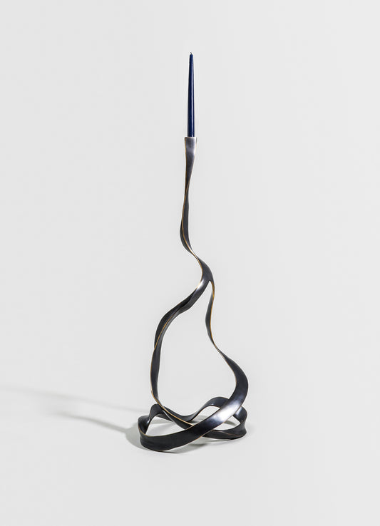 A sculptural metal candle holder with a twisted, wave-like design in a dark, matte finish, holding a tall, slender blue candle at the top. The piece has an abstract, contemporary style.