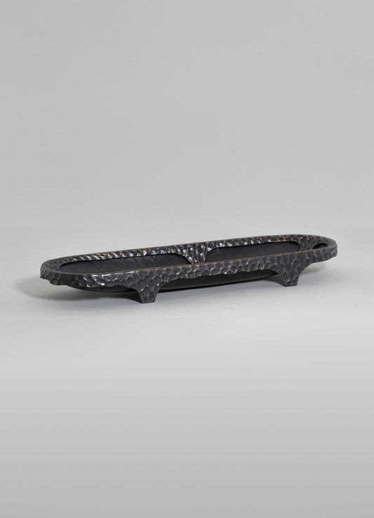 A black, hammered metal tray with an elongated oval shape, raised edges, and three short, curved legs. The tray features a textured surface with a rustic, handcrafted style.