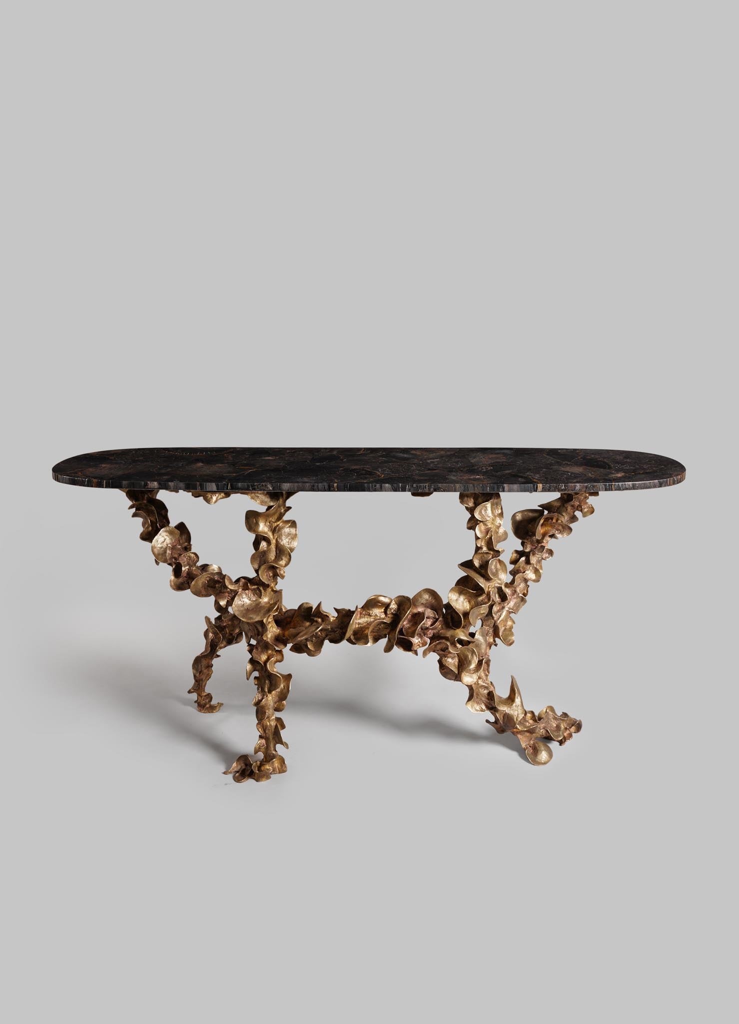 A table with a dark, polished stone top and an ornate, sculptural base made of intertwined, irregularly shaped metal in a gold finish.