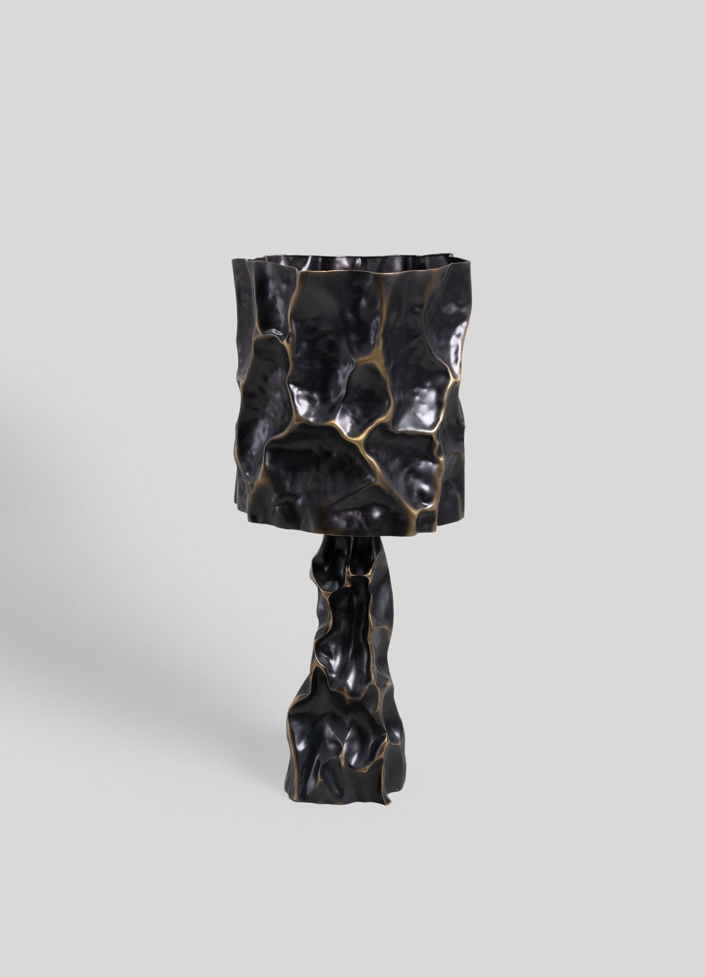 A black ceramic table lamp with an irregular, textured, and organic-shaped surface, featuring gold accents that highlight its sculptural form. The design exudes a contemporary and artisanal aesthetic with a glossy finish.