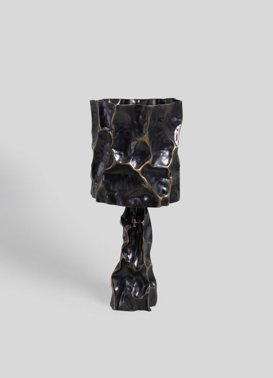 A black ceramic table lamp with an irregular, textured, and organic-shaped surface, featuring gold accents that highlight its sculptural form. The design exudes a contemporary and artisanal aesthetic with a glossy finish.