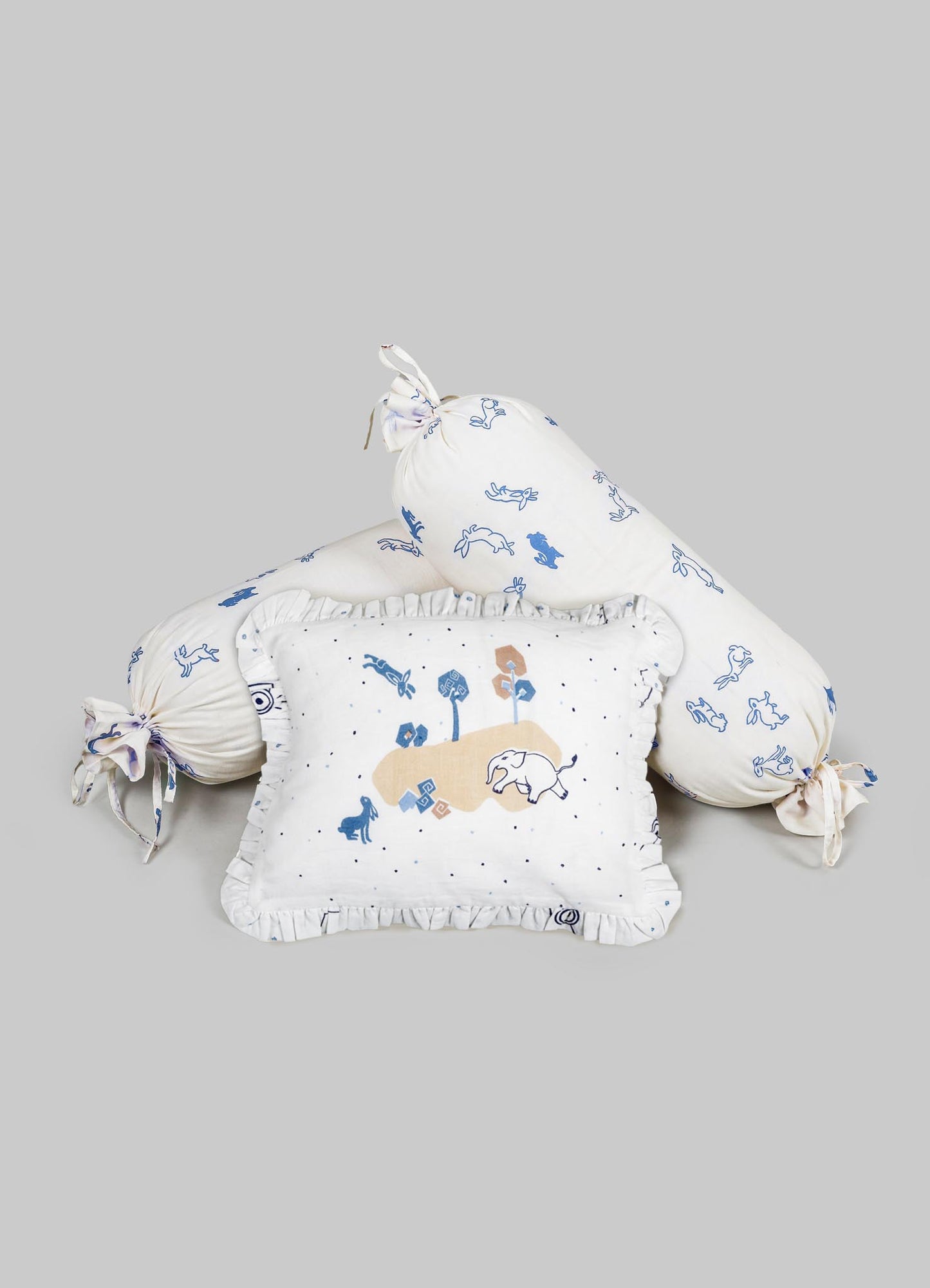 Cotton bedding set with printed animal motifs in blue and beige, including a square cushion and two cylindrical bolster pillows with tied ends. The fabric features playful illustrations and ruffled trim details.