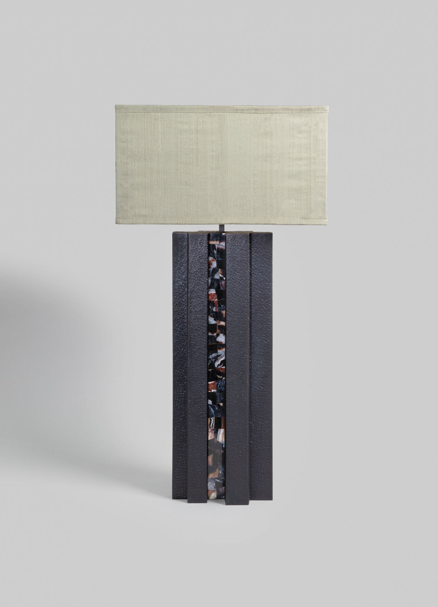 A table lamp with a rectangular fabric shade in a neutral beige tone and a textured black base featuring a vertical decorative panel with a multicolored mosaic pattern.
