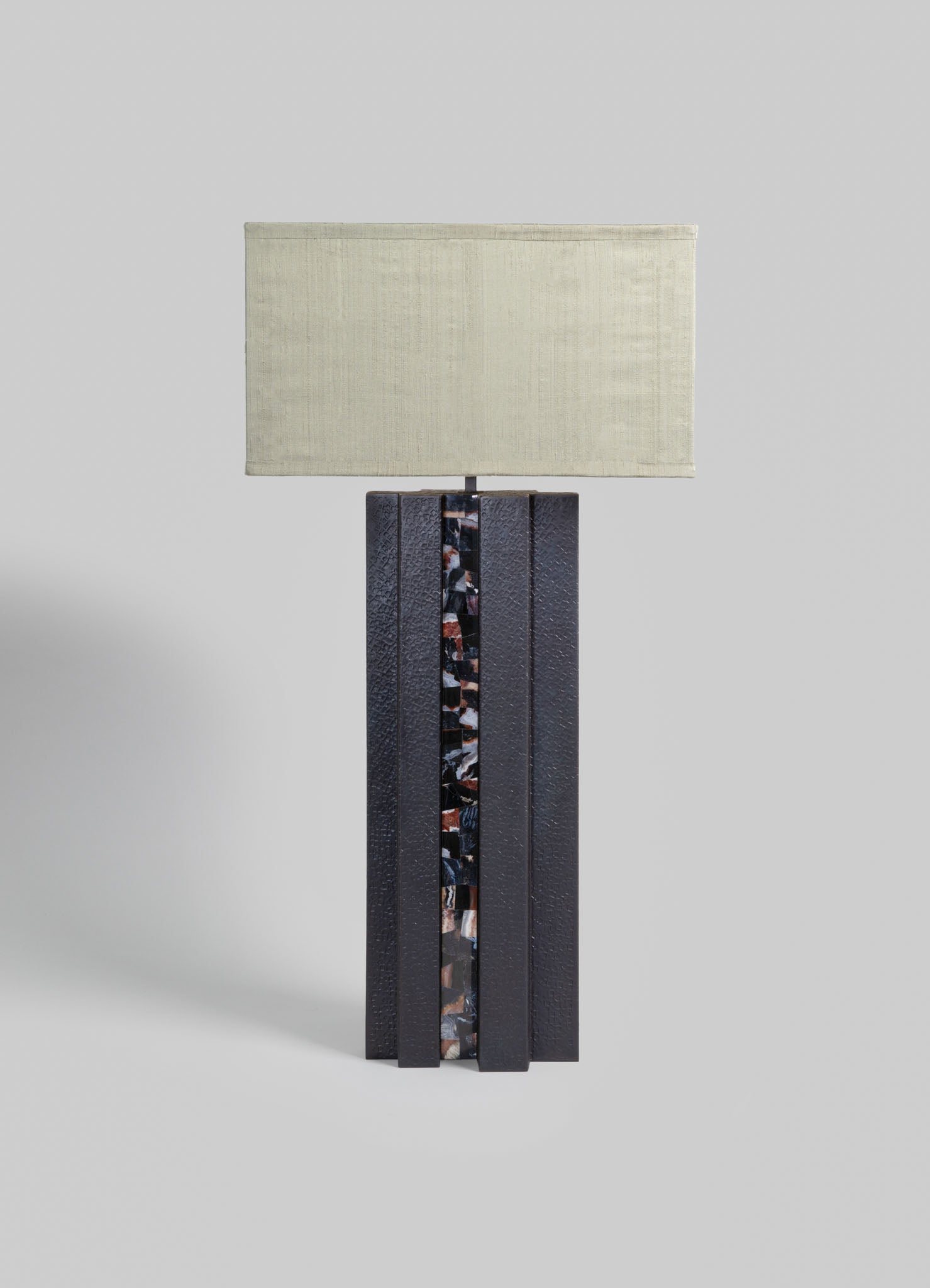 A table lamp with a rectangular fabric shade in a neutral beige tone and a textured black base featuring a vertical decorative panel with a multicolored mosaic pattern.