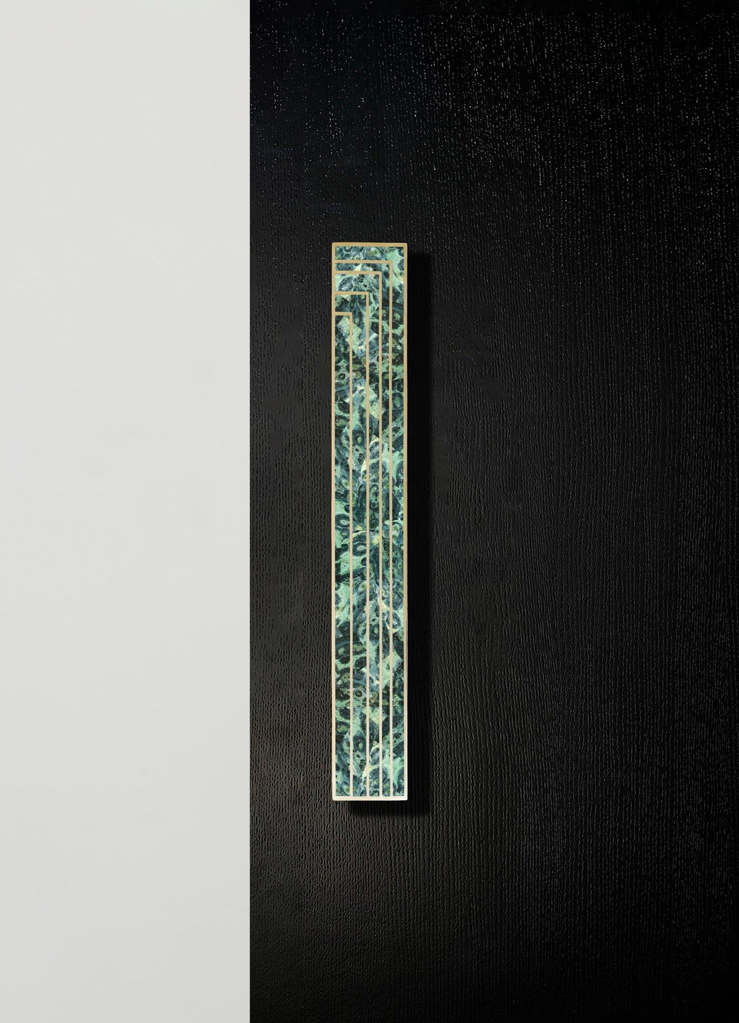 A narrow decorative panel with a botanical motif in shades of green and blue, with a gold frame, mounted vertically on a wall with contrasting black and white sections.