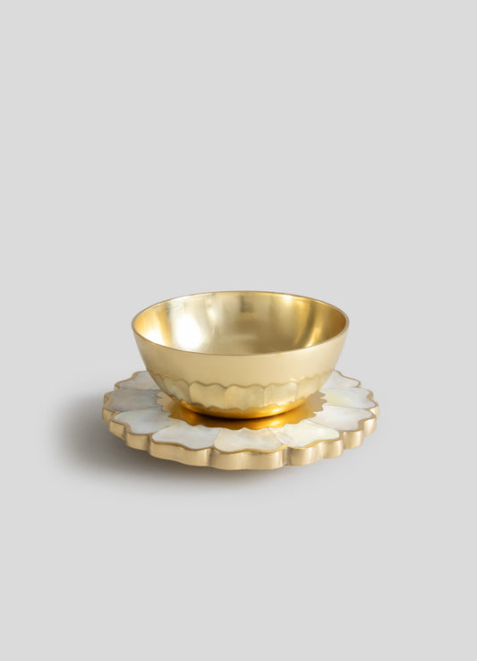 A metallic gold bowl with a smooth, rounded shape sits on a scalloped, flower-shaped base made of mother-of-pearl or similar material. The design combines elegant, luxury materials with a contemporary style.