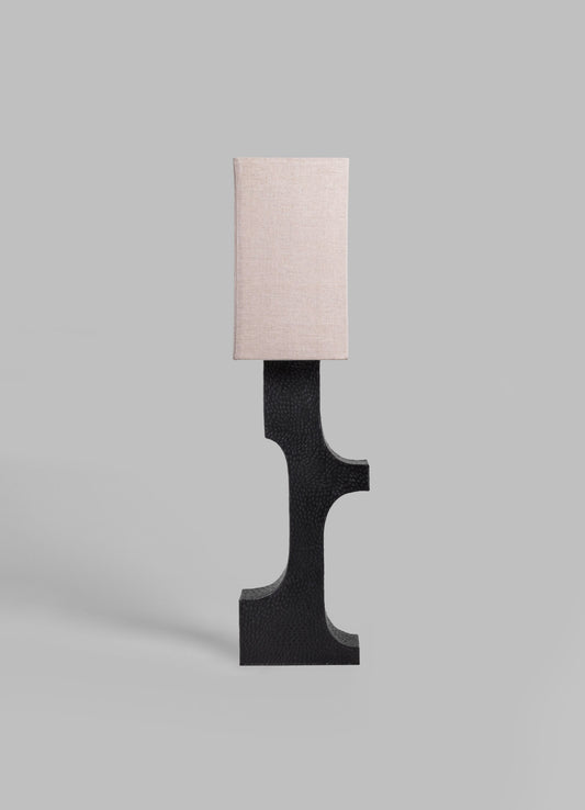 A table lamp with a rectangular beige fabric lampshade and a black textured base with curved and geometric shapes, set against a neutral background.
