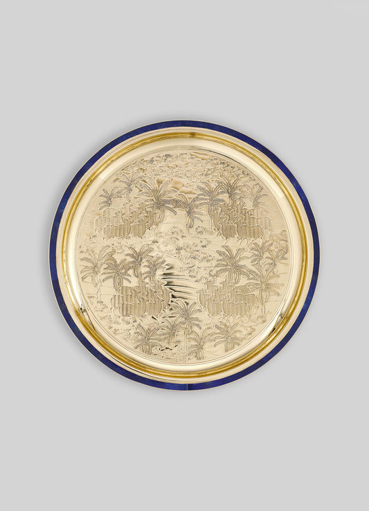 A decorative round tray with an etched tropical landscape featuring palm trees and a river, made of polished metal with a blue and gold border. The design has a detailed, engraved pattern creating a sophisticated, ornamental look.