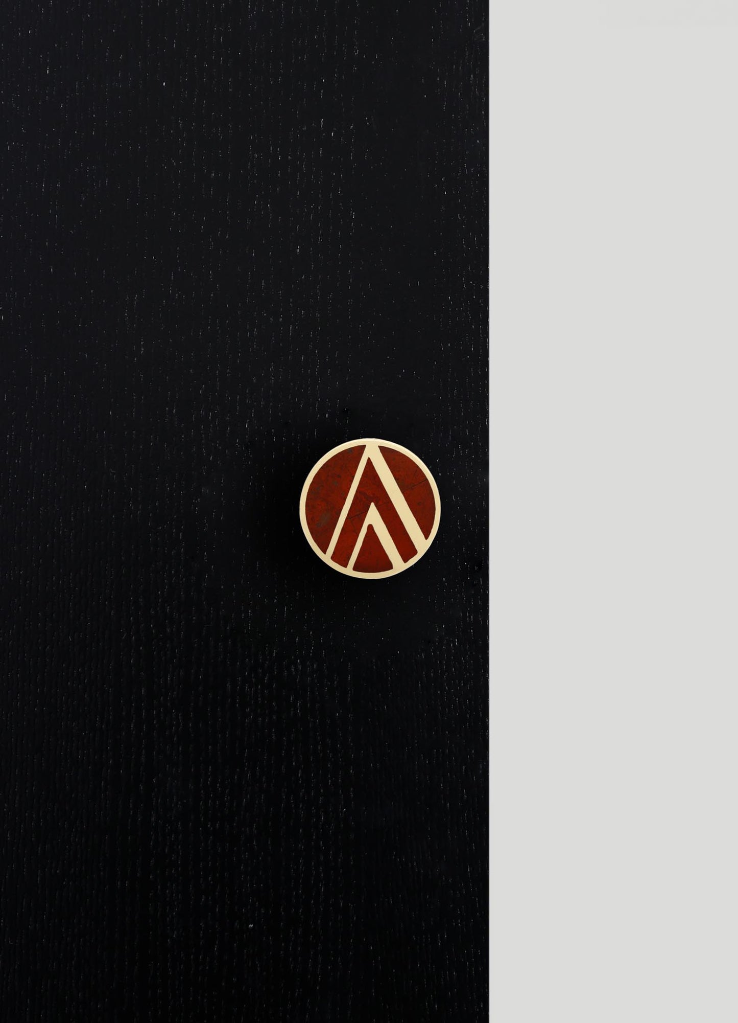 A black leather item with a brass round pin featuring a geometric pattern of upward-pointing arrows in dark red and cream tones. The background includes a light gray surface, highlighting the textures and metallic accents of the product.