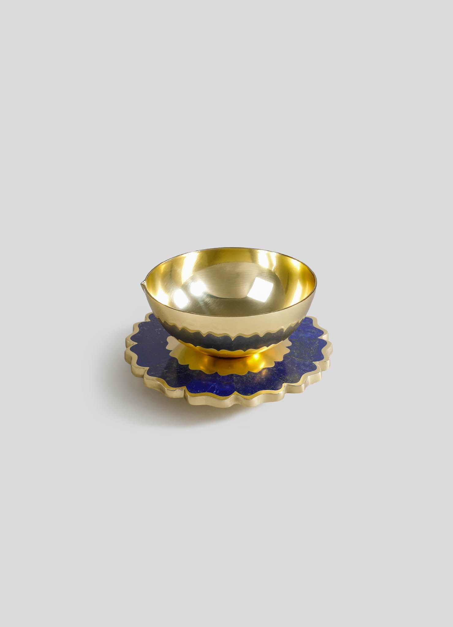 A gold-colored metal bowl with a polished, reflective surface sitting on a decorative blue and gold scalloped tray.