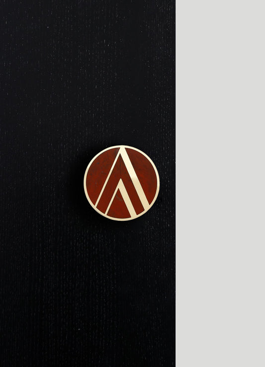 A round brooch with a geometric pattern in white and reddish-brown tones, displayed on a black textured fabric background with a light grey section on the right.