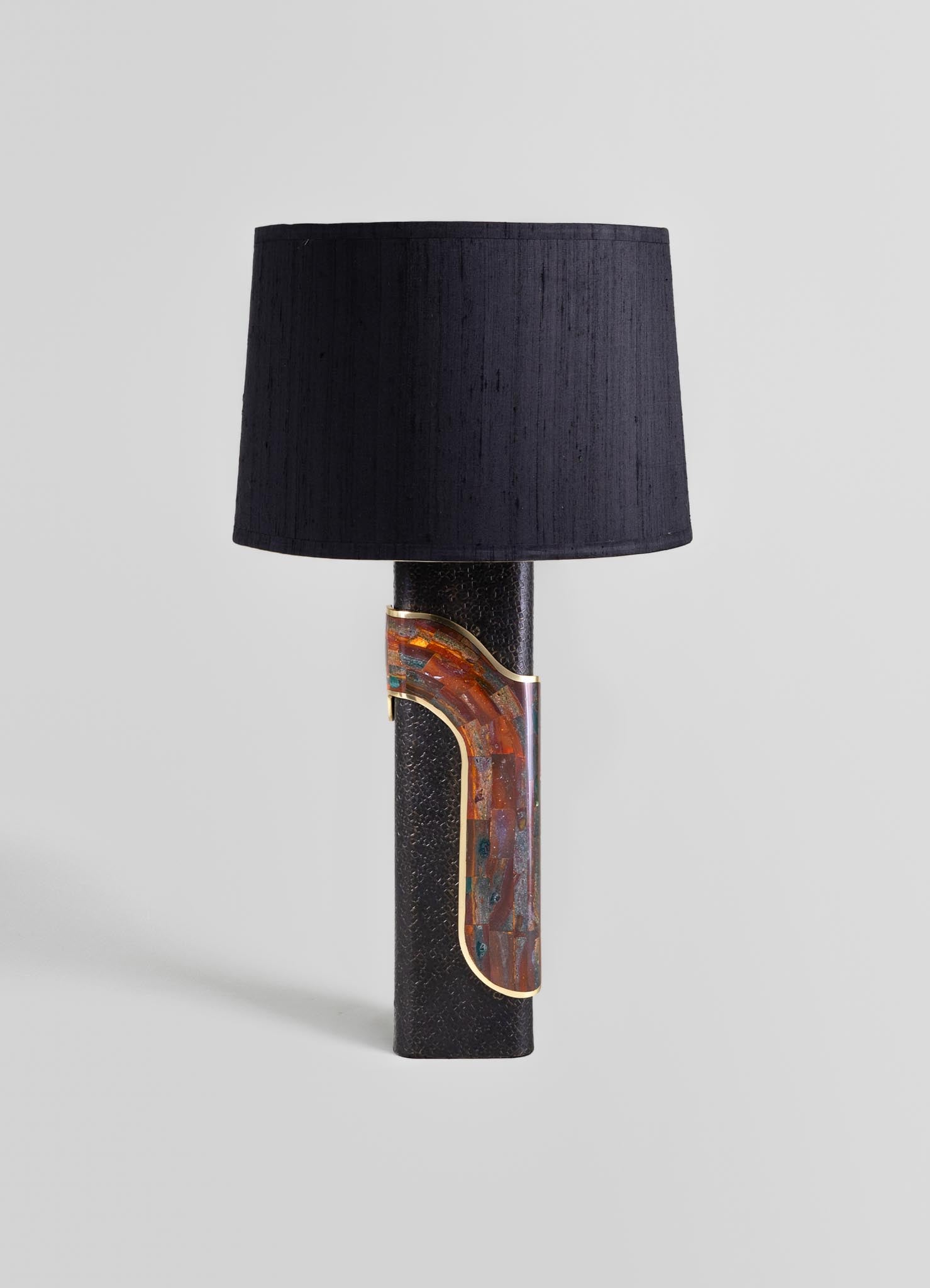 A table lamp with a textured black ceramic base featuring an abstract, curved inlay with warm earthy tones and metallic accents, topped with a large black fabric drum shade.