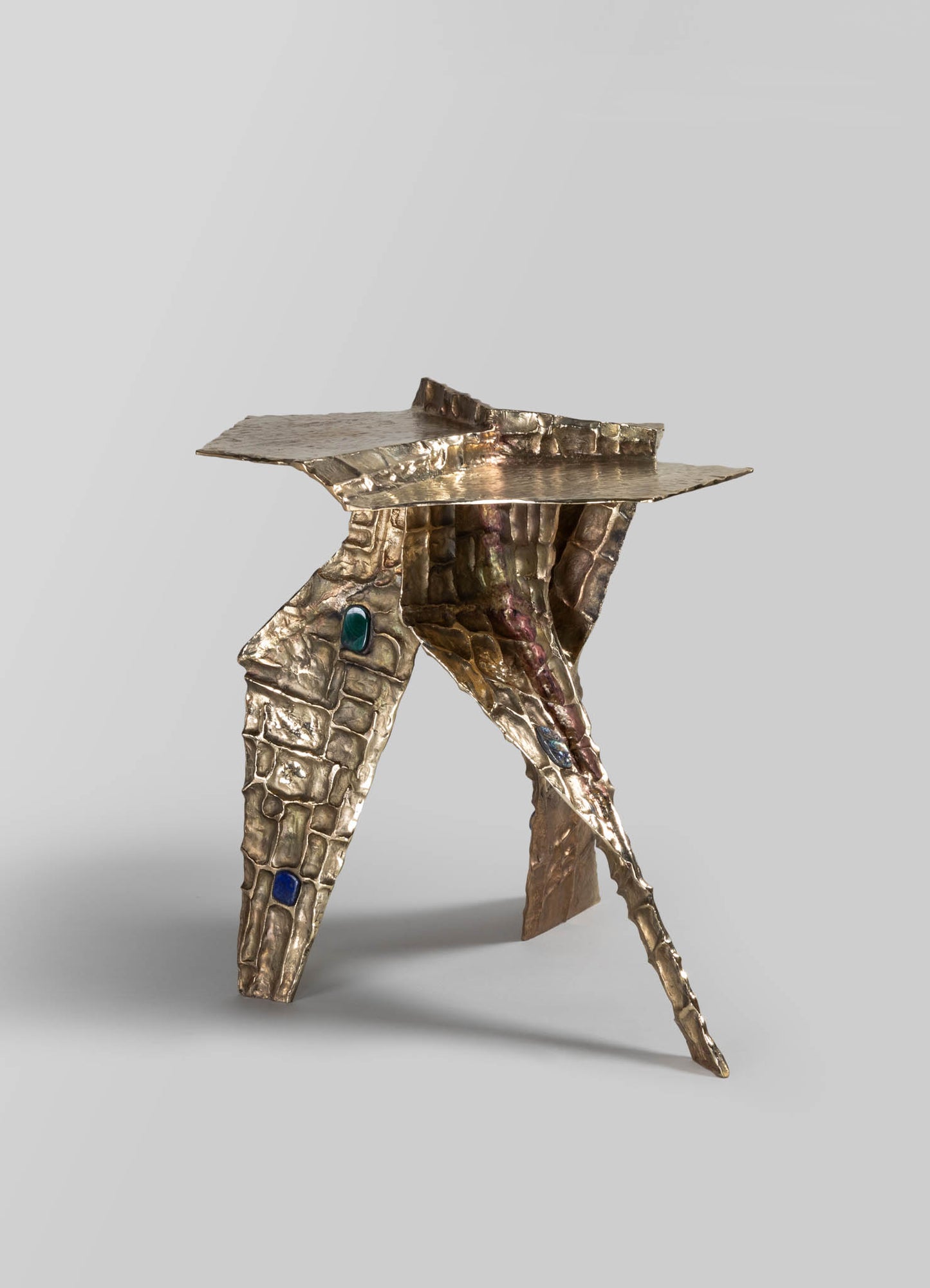 A metallic sculpture with a hammered texture and an abstract form, featuring embedded colored stones or glass elements in shades of blue and green. The piece has an organic, handcrafted appearance with a mix of shiny metal finish.