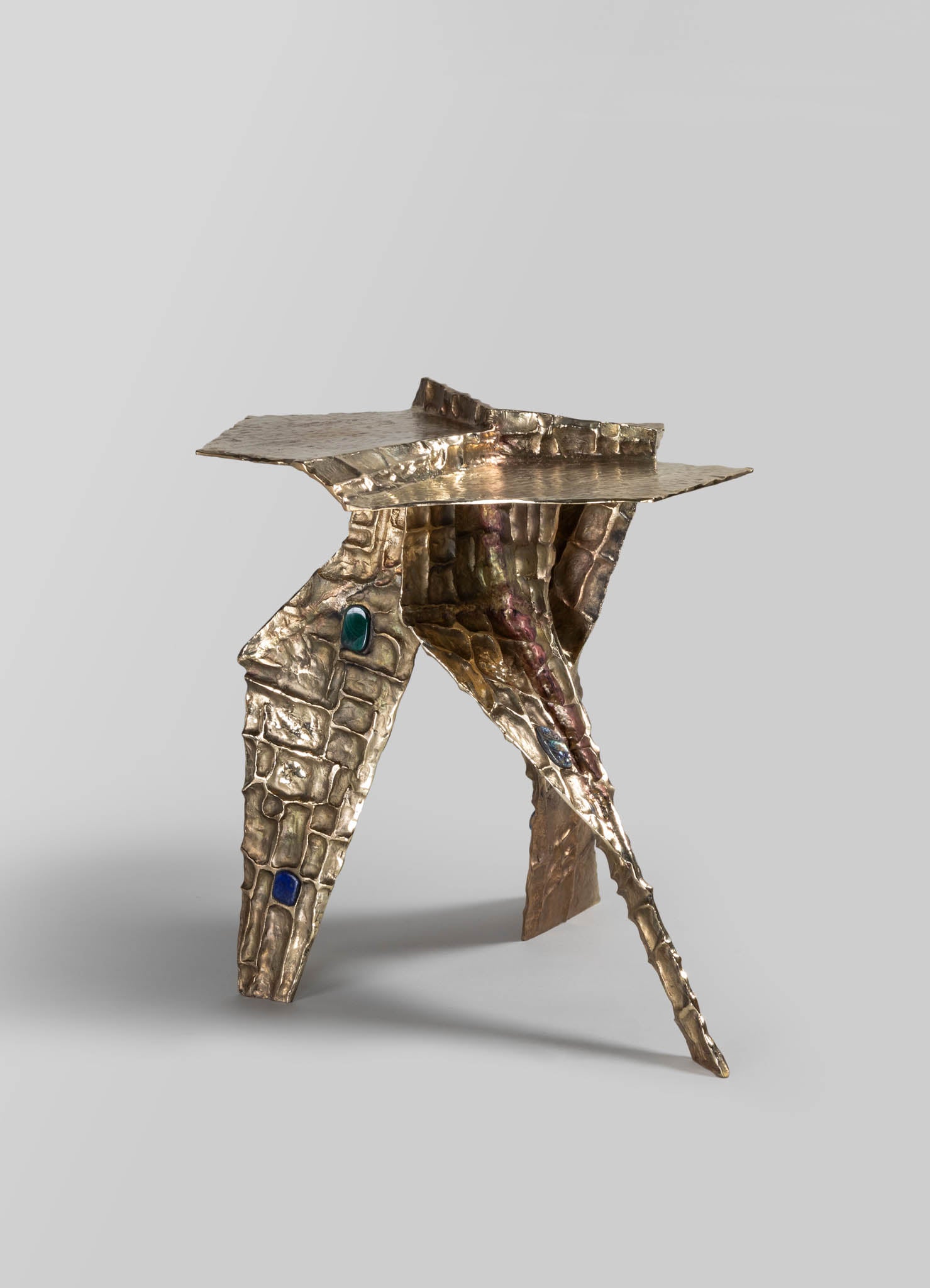 A metallic sculpture with a hammered texture and an abstract form, featuring embedded colored stones or glass elements in shades of blue and green. The piece has an organic, handcrafted appearance with a mix of shiny metal finish.
