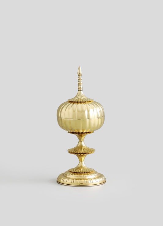 A decorative gold-colored container with a rounded, fluted body, mounted on a tiered base with ribbed details, topped with a pointed finial and a removable lid. The design features ornate, classical motifs with a polished metallic finish.