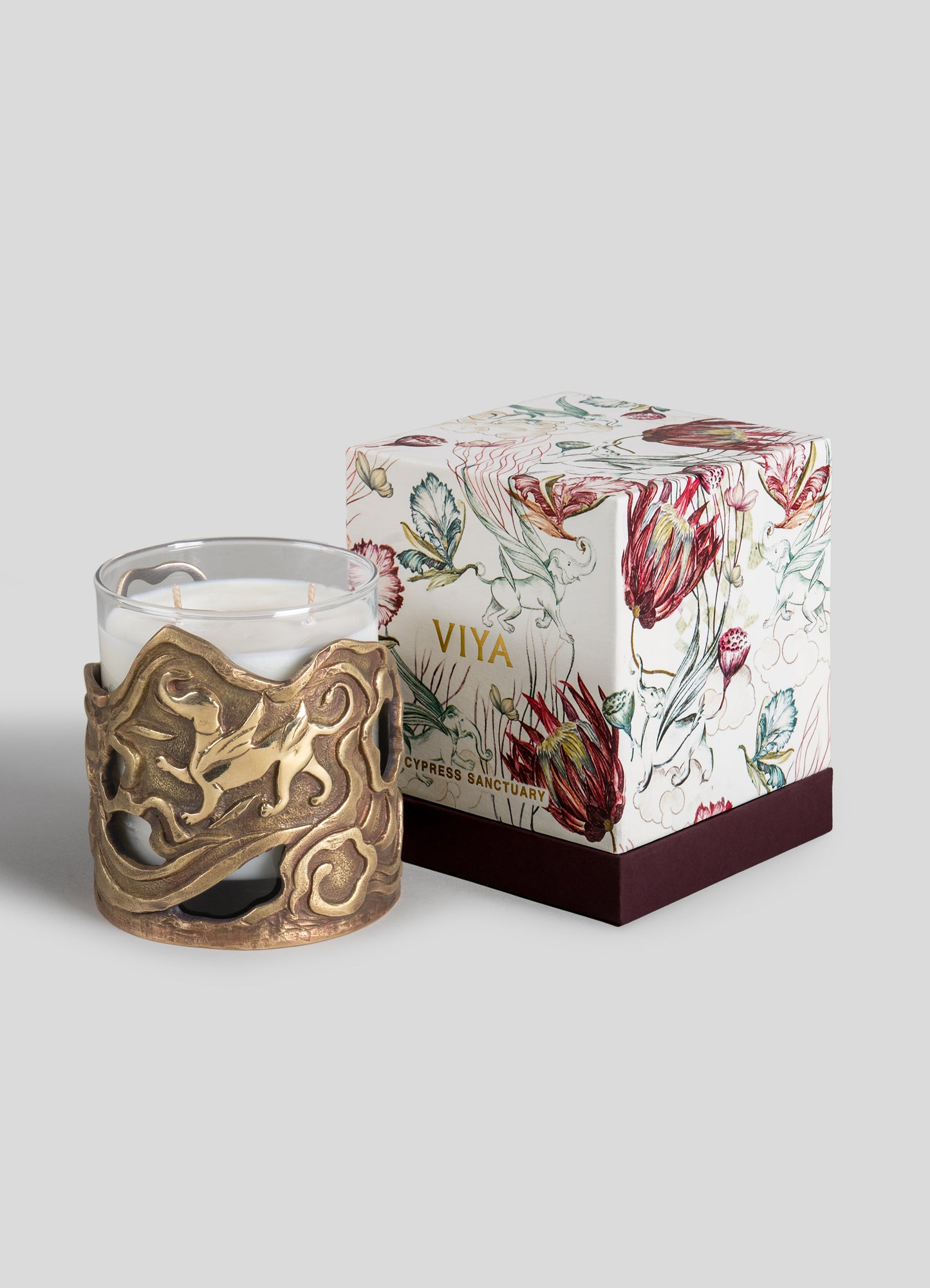 A white scented candle in a decorative brass holder with an intricate, organic motif, accompanied by a floral-illustrated gift box with the label "VIYA" and "Cypress Sanctuary."