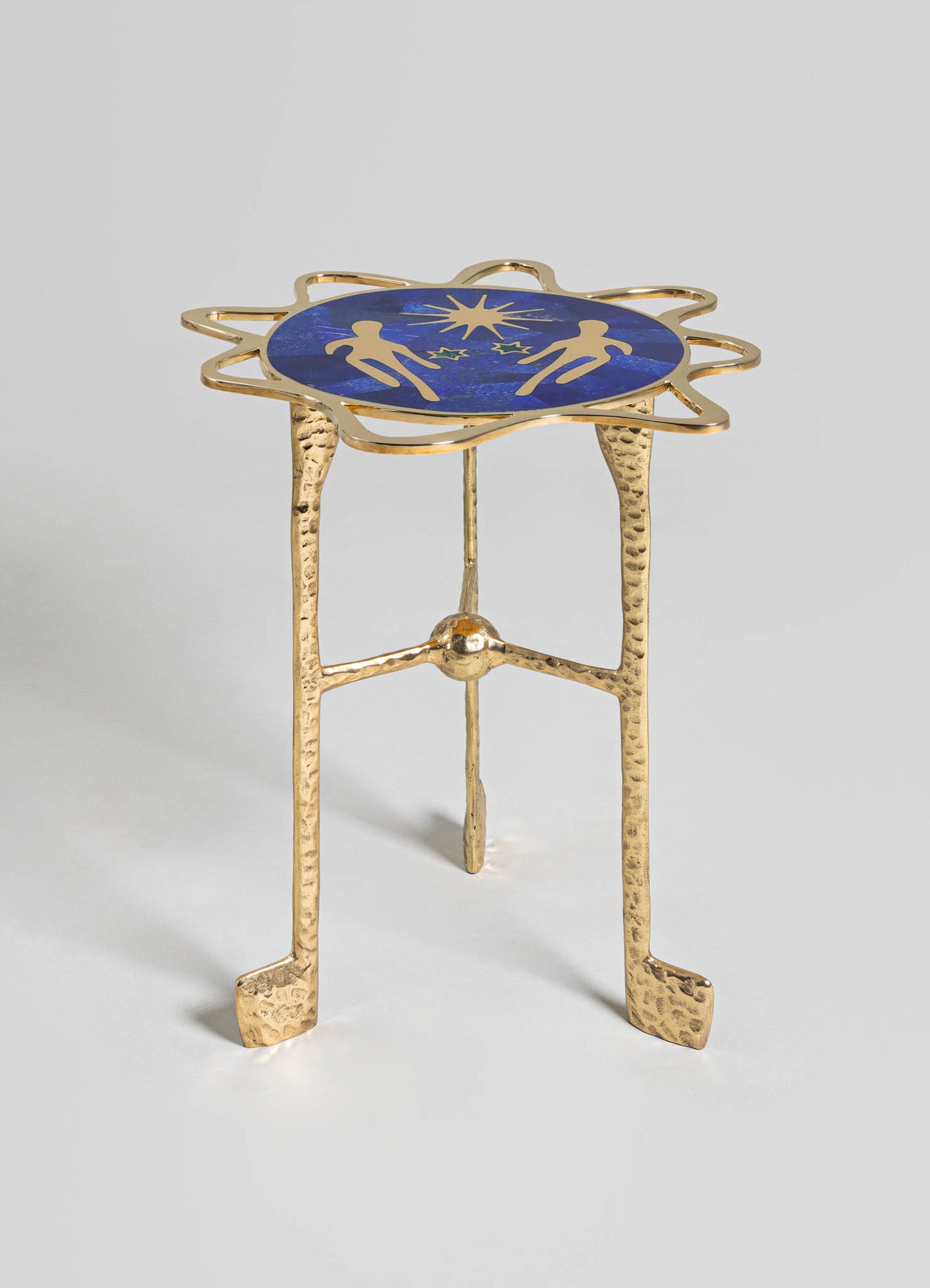 A small side table with a round top featuring a blue enamel surface decorated with gold silhouette motifs of figures and a starburst pattern. The table has a sculptural, hammered gold-tone metal frame with three legs and a decorative, irregularly shaped border around the top.