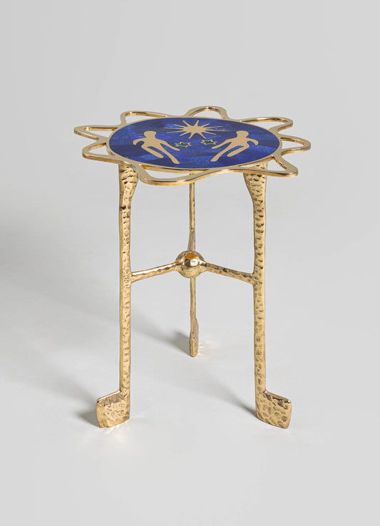 A small side table with a round top featuring a blue enamel surface decorated with gold silhouette motifs of figures and a starburst pattern. The table has a sculptural, hammered gold-tone metal frame with three legs and a decorative, irregularly shaped border around the top.