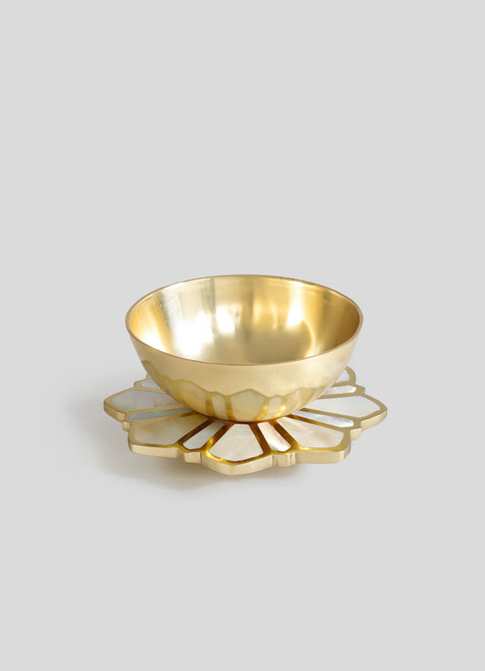 A brass bowl with a polished, reflective finish placed on a decorative mother-of-pearl and brass scalloped tray, featuring a floral motif and a combination of metallic and iridescent tones.