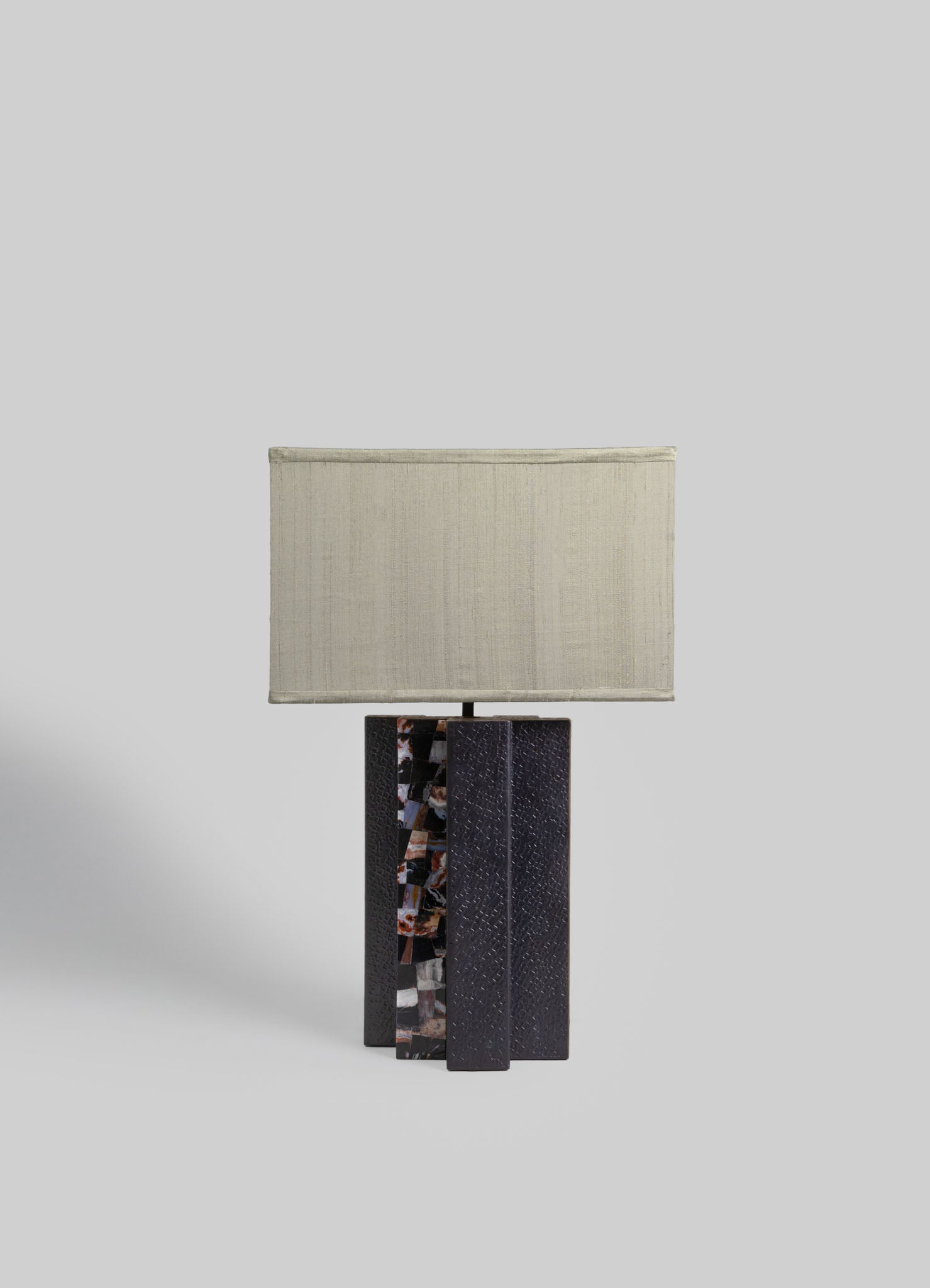 A table lamp with a rectangular fabric shade in a neutral beige tone, mounted on a base composed of three vertical textured black materials with one featuring a multicolored mosaic pattern.