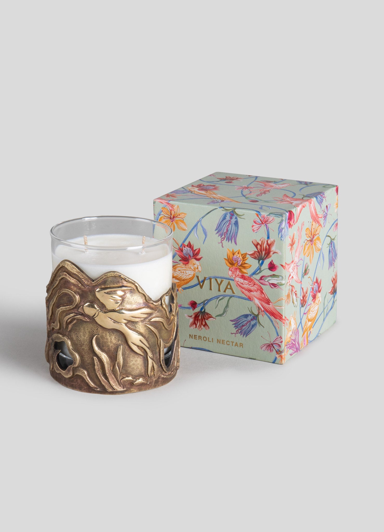 A white scented candle in a glass holder with a brass-like decorative metal sleeve featuring abstract organic shapes. It is paired with a floral-themed gift box with colorful botanical illustrations and the label "Viya Neroli Nectar."