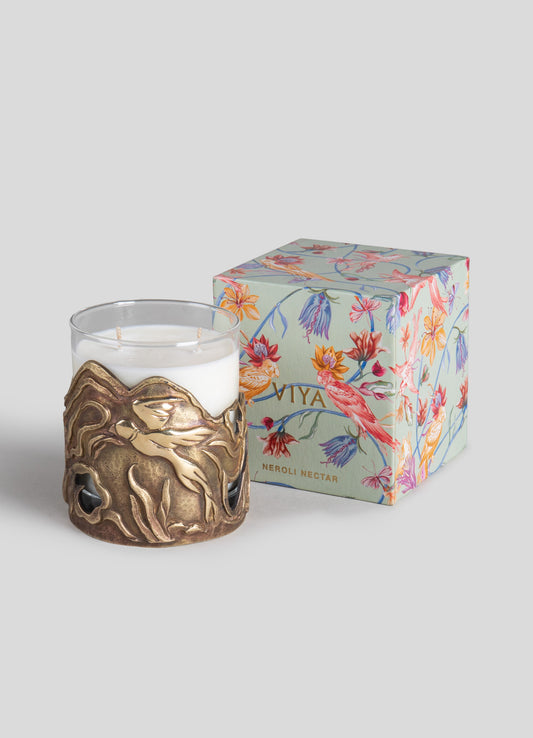 A white scented candle in a glass holder with a brass-like decorative metal sleeve featuring abstract organic shapes. It is paired with a floral-themed gift box with colorful botanical illustrations and the label "Viya Neroli Nectar."