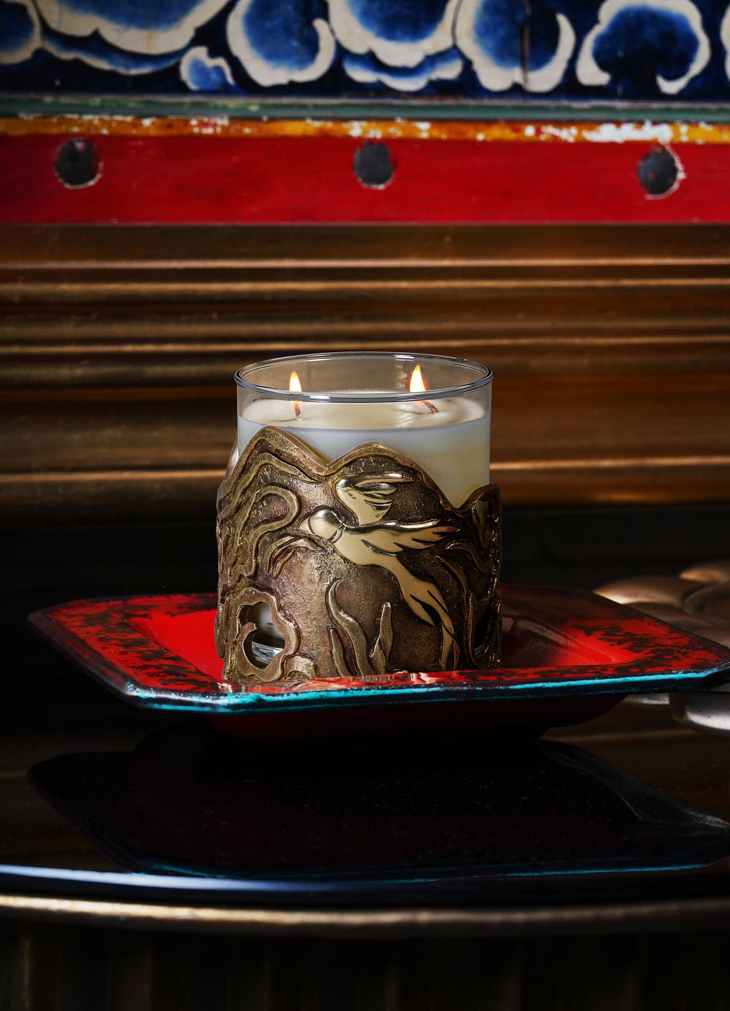 A glass candle holder with a metallic gold finish featuring an intricate dragon motif, containing a lit white candle. It is placed on a red and black ceramic tray with a distressed finish.