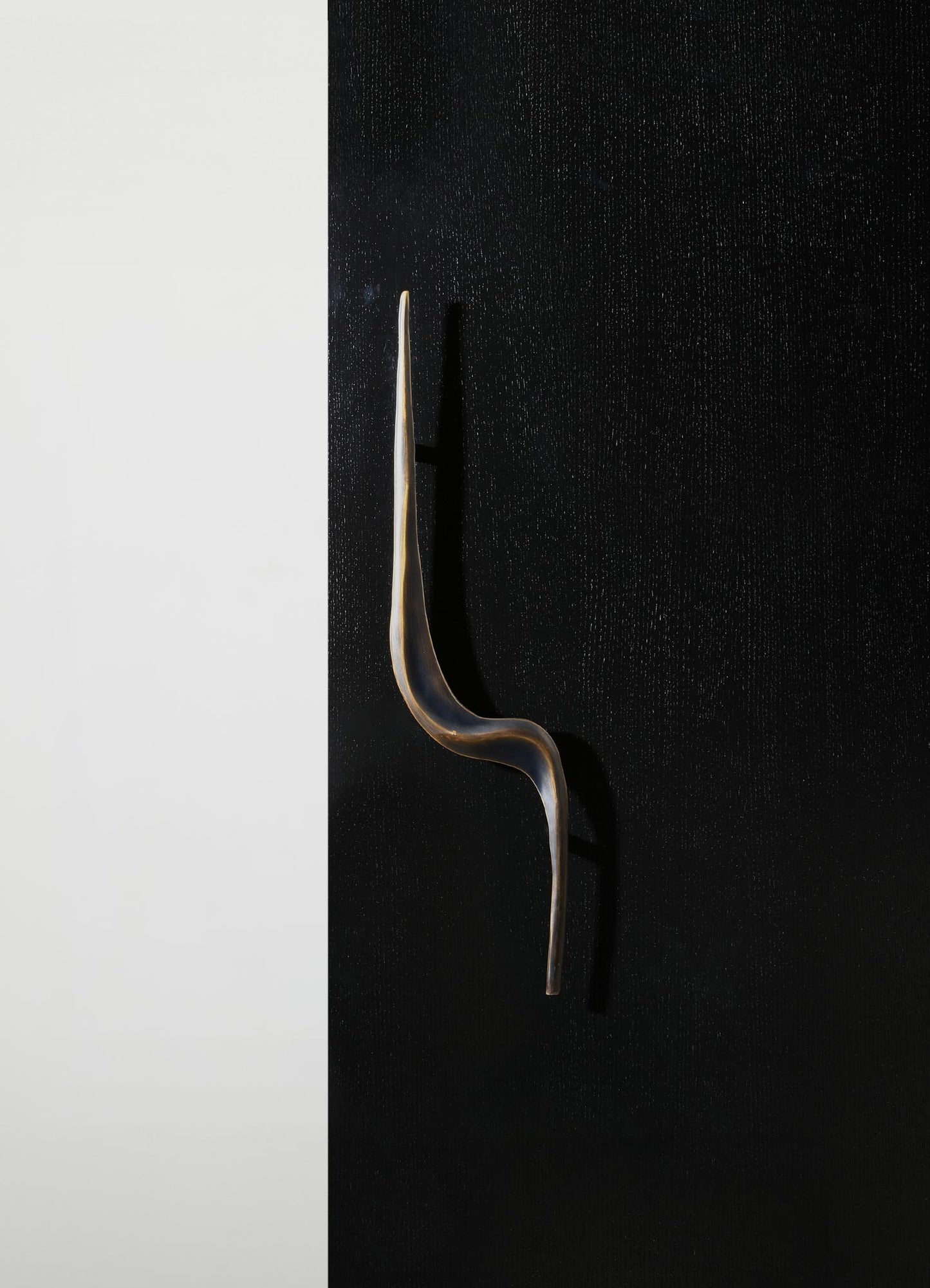 A sleek, modern door handle with a curved, elongated shape in brushed metal finish mounted on a black textured door, contrasting with a smooth white wall in the background.