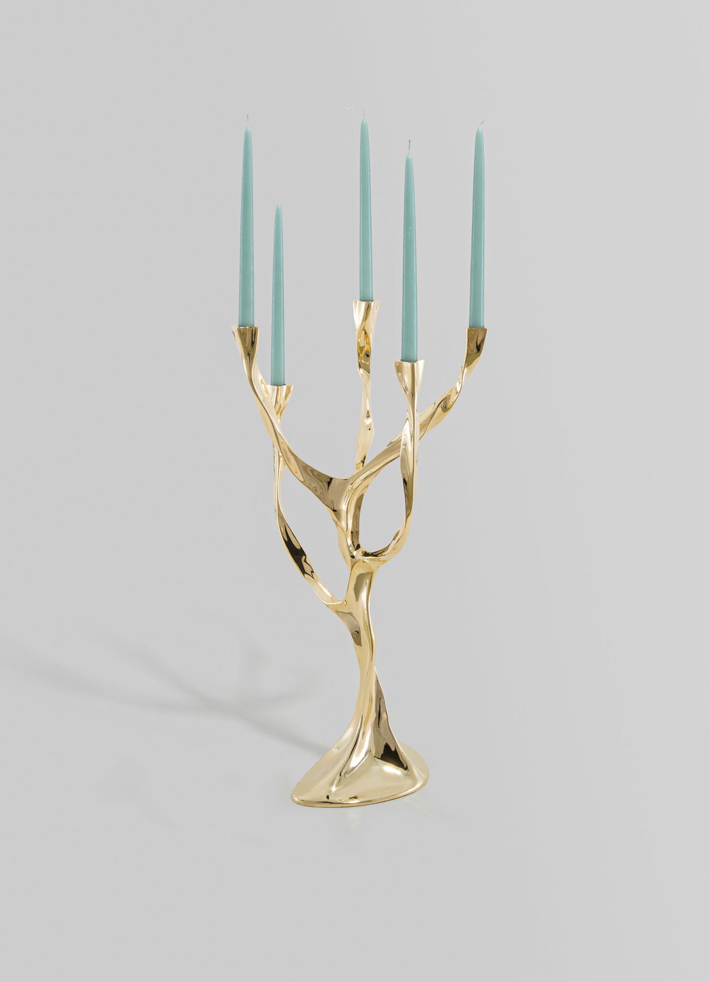 A brass candlestick holder with an abstract, sculptural design resembling intertwined branches, holding six light blue taper candles. The piece has a polished gold finish and a contemporary style.
