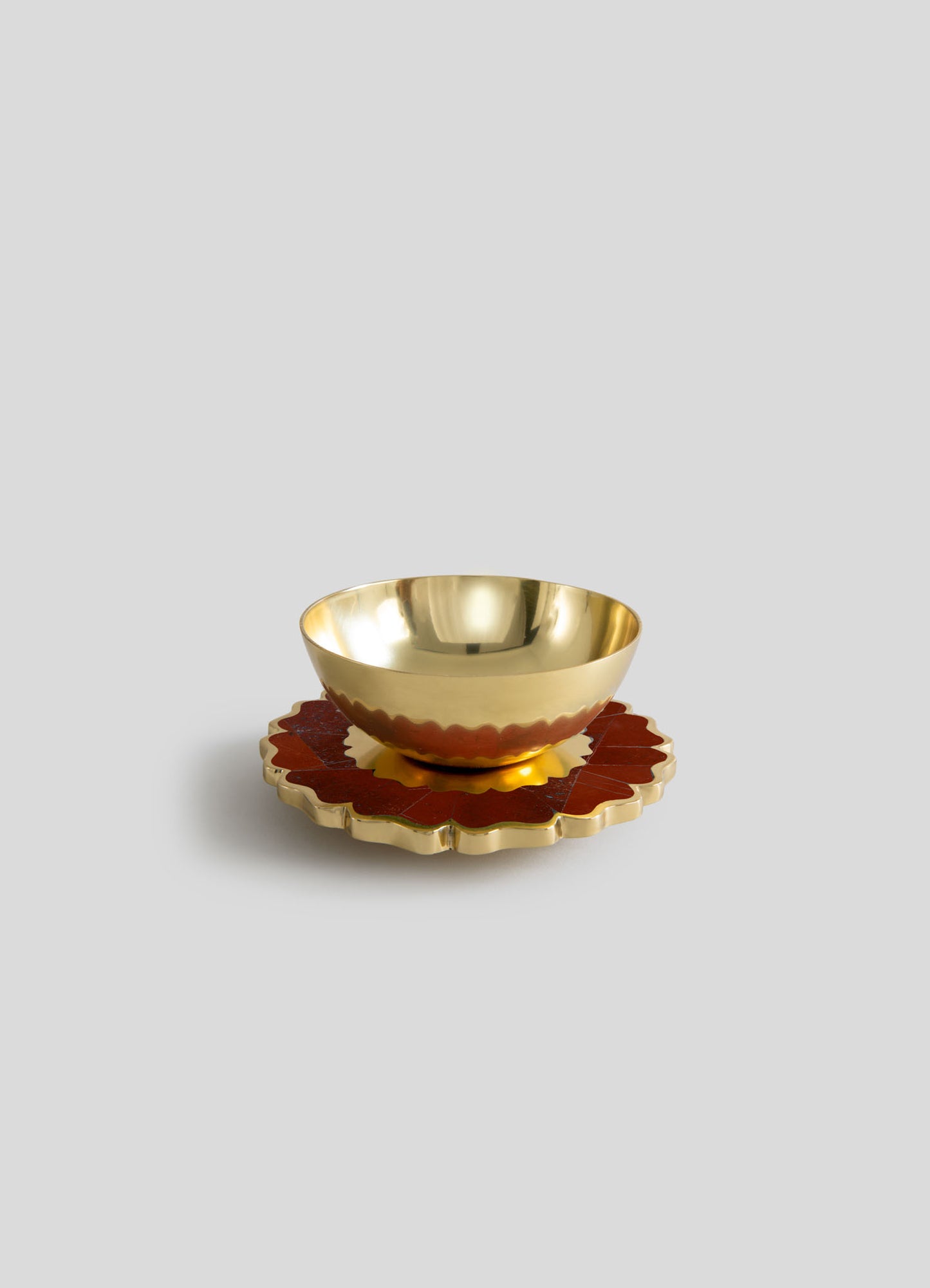 A decorative bowl with a polished gold interior and a scalloped edge, placed on a matching scalloped base with a reddish-brown finish. The set features metallic and wooden materials with an ornate, handcrafted style.