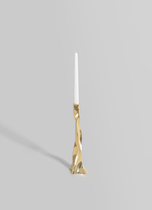 A sculptural candle holder made of metal with a polished gold finish, featuring an irregular, fluid shape. It holds a tall, white taper candle and has a contemporary, artistic style.