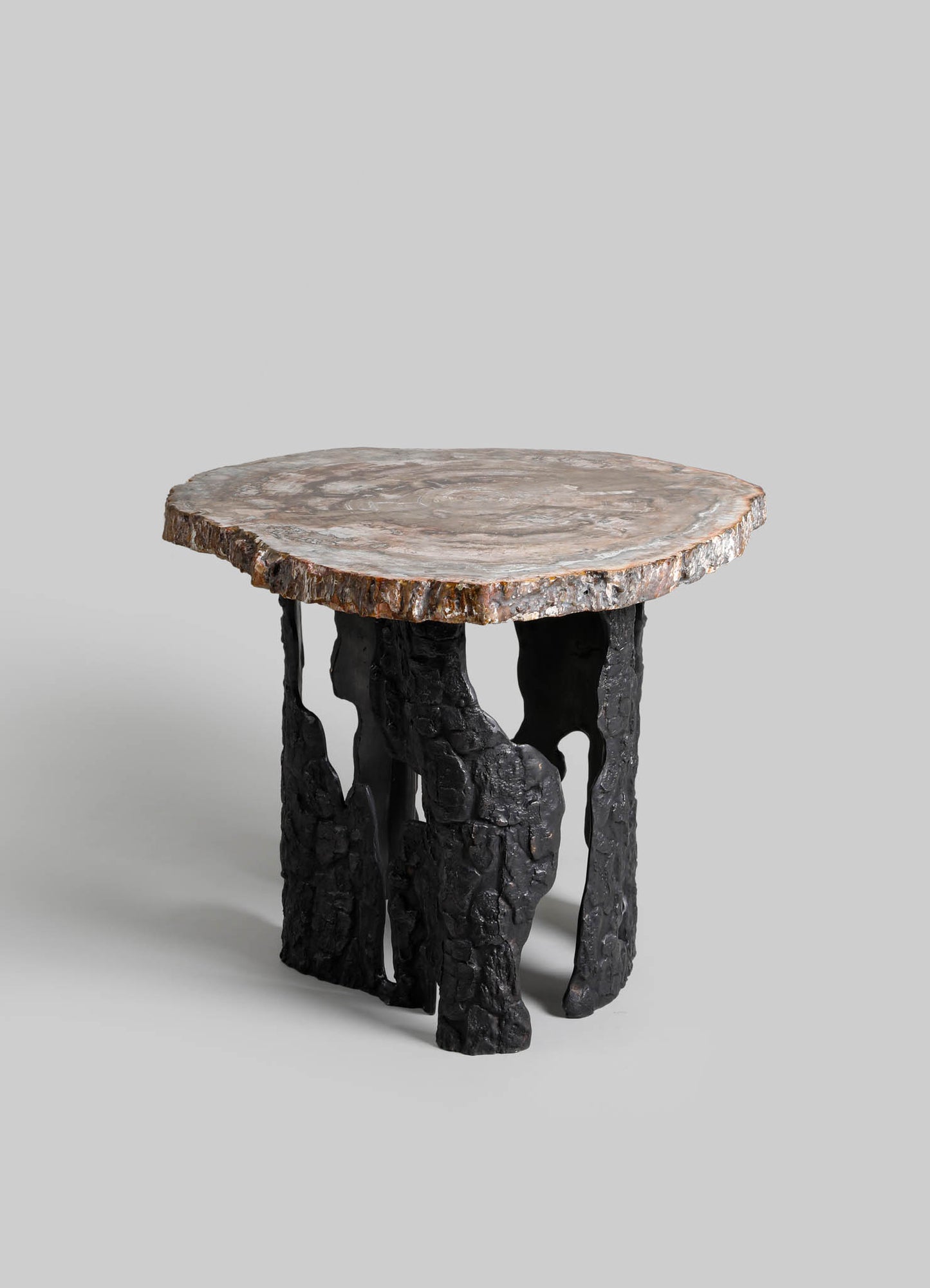 Round side table with a natural stone top displaying beige, gray, and white tones, supported by black, textured, irregularly shaped legs resembling driftwood or sculpted wood.