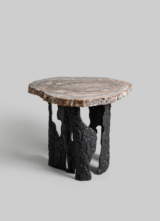Round side table with a natural stone top displaying beige, gray, and white tones, supported by black, textured, irregularly shaped legs resembling driftwood or sculpted wood.