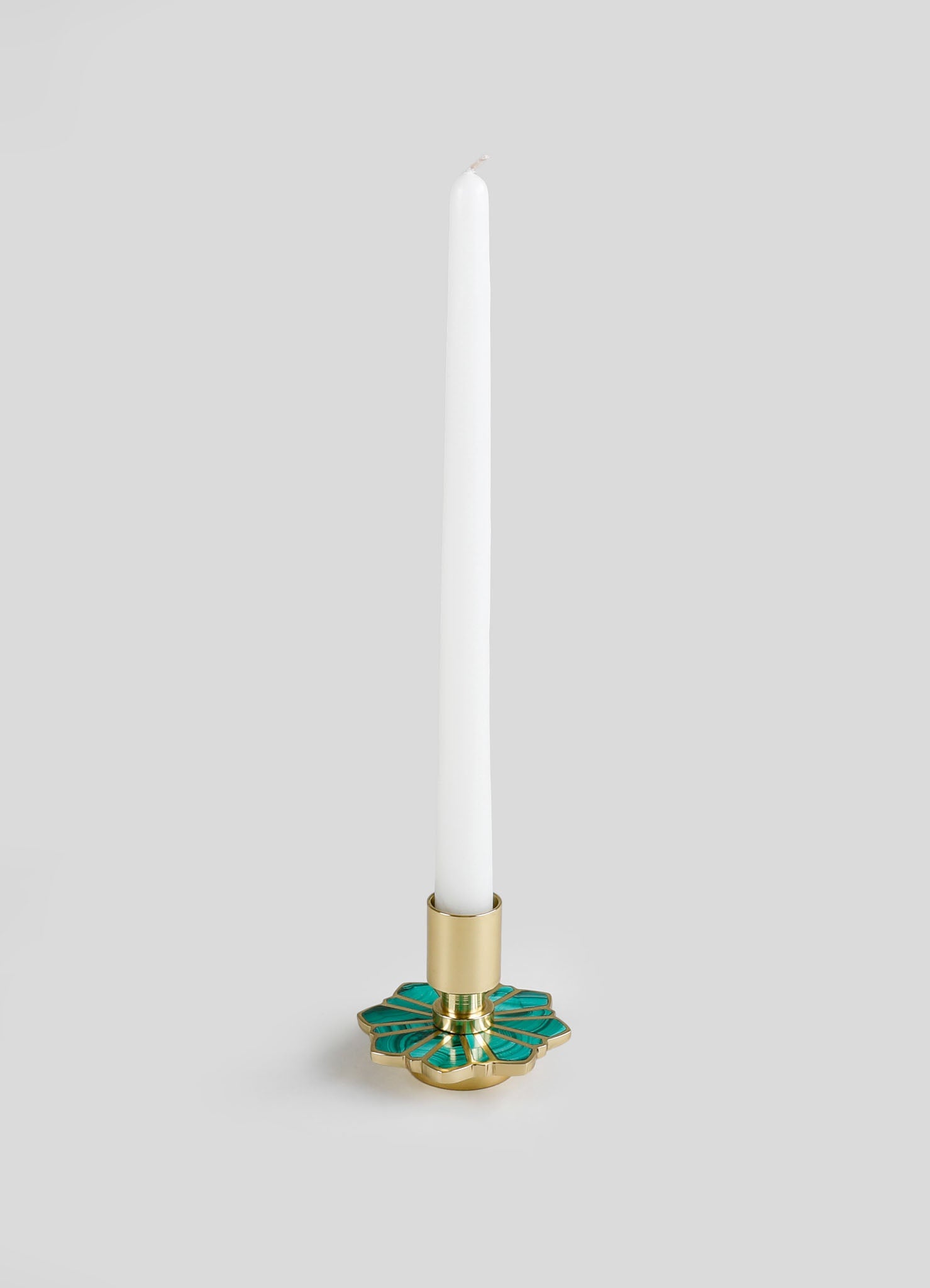 A tall white candle in a decorative brass candle holder with green enamel accents, featuring a geometric floral motif.