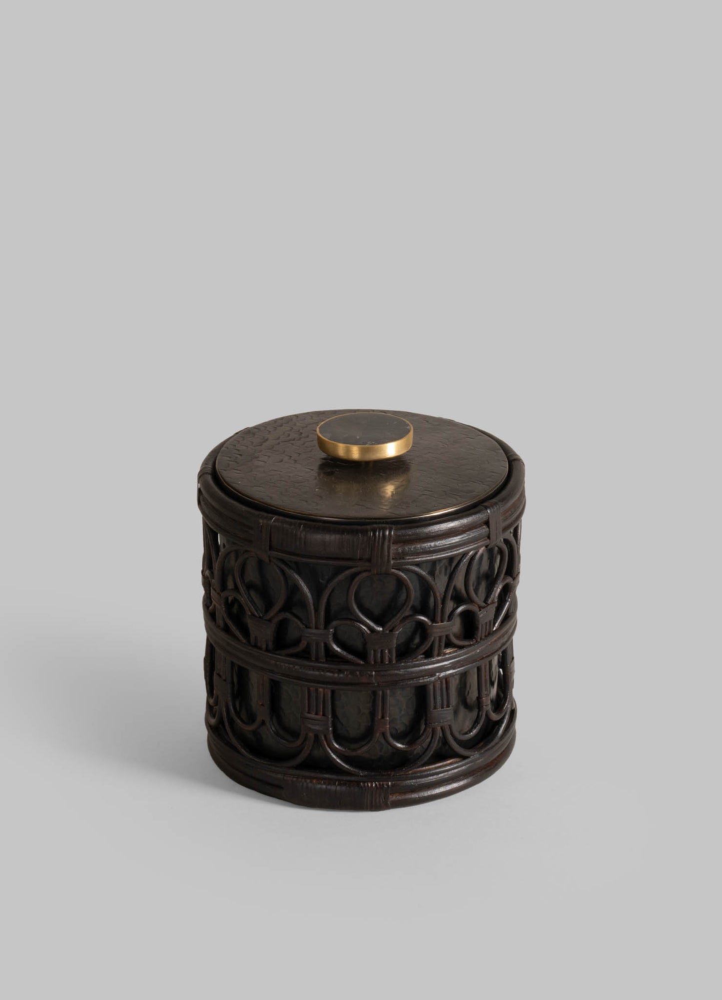 The VIYA Manipur Ice Bucket Small is a cylindrical, dark wood container with intricate carvings and a flat lid topped by a round, hand-beaten brass knob, shown against a plain light gray background.