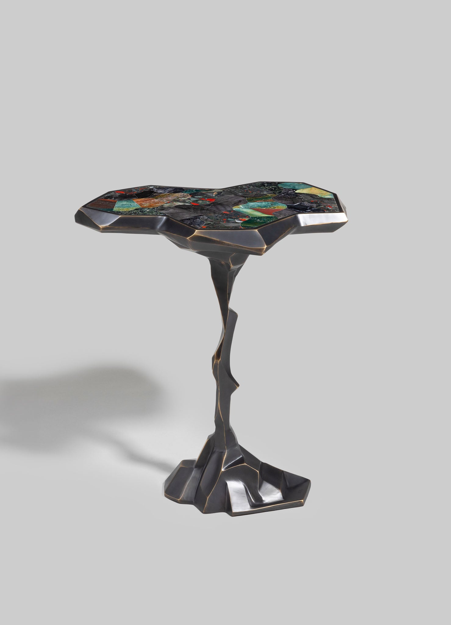 A decorative side table with an irregular-shaped tabletop made of colorful mosaic tiles in green, red, black, and blue tones, set on a sculptural black metal base with gold accents.