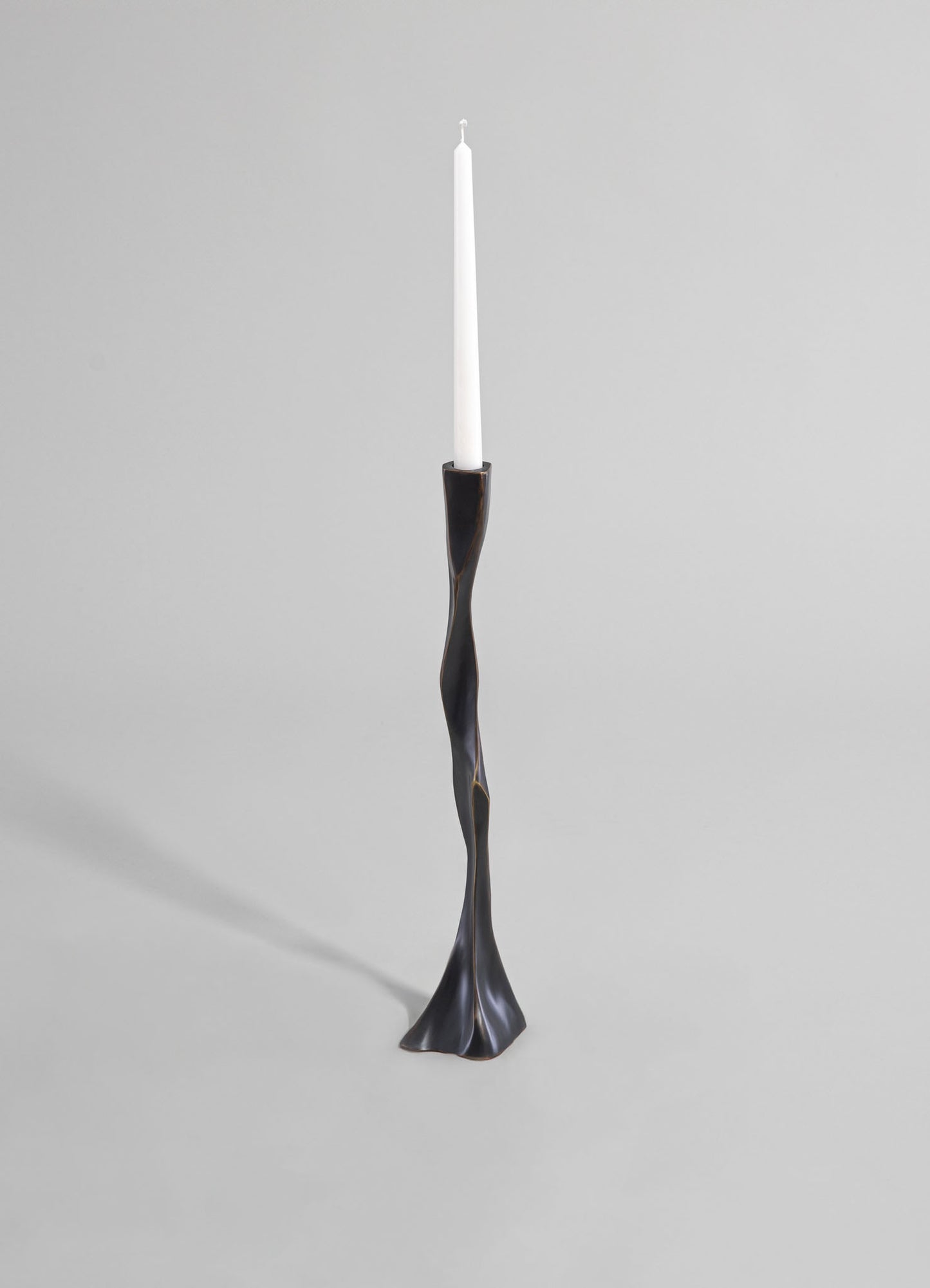 A tall, sculptural candle holder with a twisted, organic shape in dark metal or black finish, holding a white taper candle. The design features a fluid, contemporary style with a smooth surface and a wide, textured base.