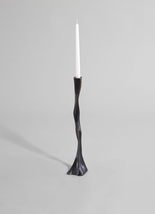 A tall, sculptural candle holder with a twisted, organic shape in dark metal or black finish, holding a white taper candle. The design features a fluid, contemporary style with a smooth surface and a wide, textured base.