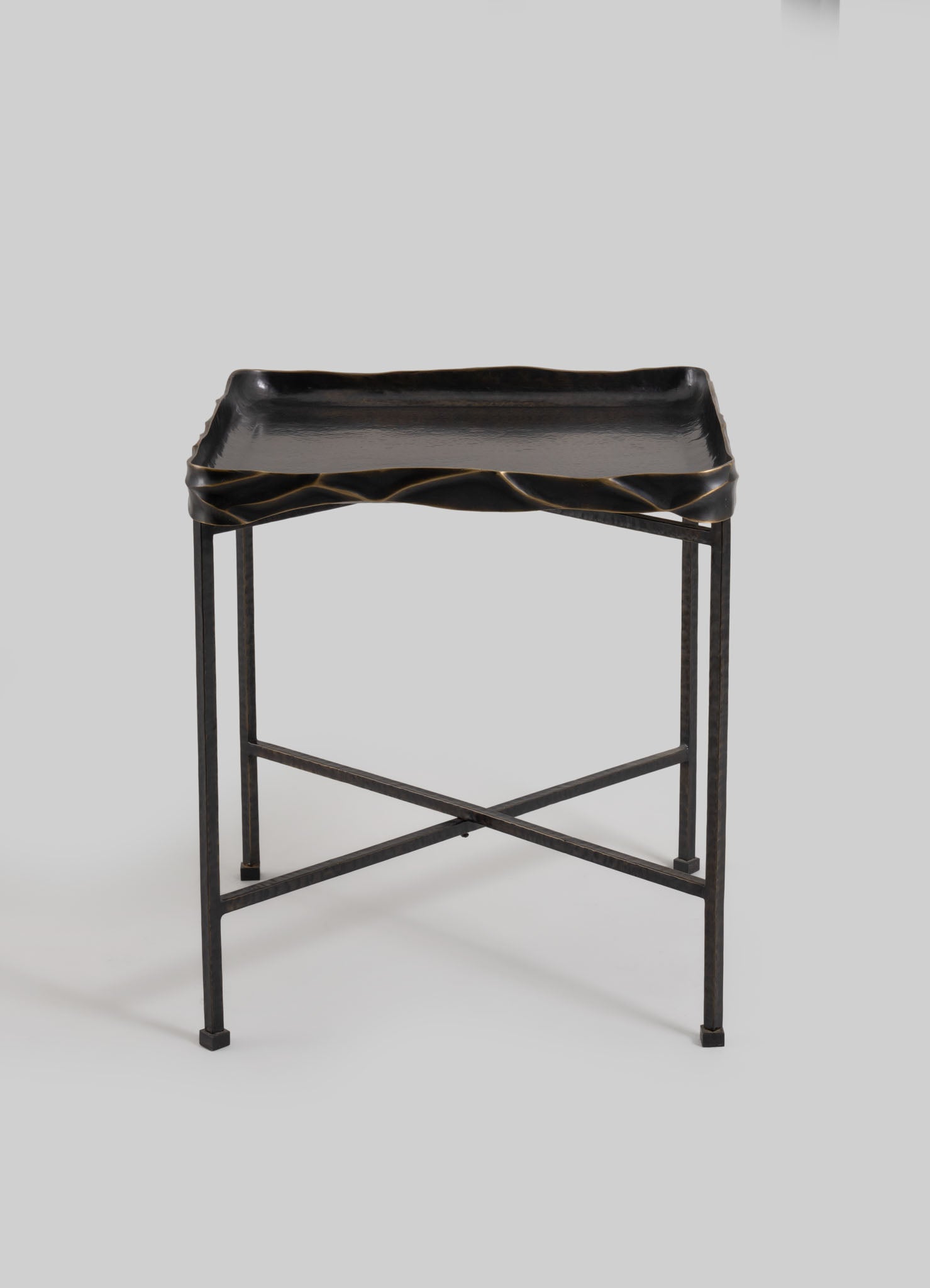 A black metal side table with a rectangular tray top featuring a slightly wavy, textured edge. The table has a minimalist frame with crossed supports and small square feet, emphasizing modern and industrial design.